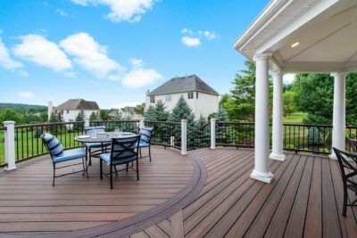 Wooden deck with table and chairs, next to a covered porch. Green trees and houses in the background.